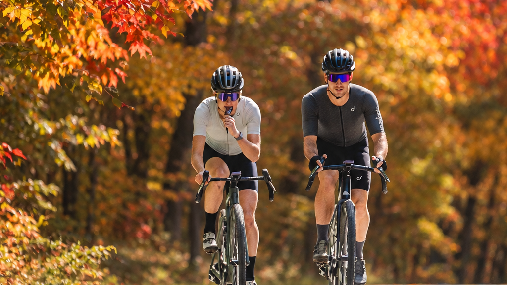 Two cyclists riding on an autumn-lined road with vibrant orange and yellow foliage.