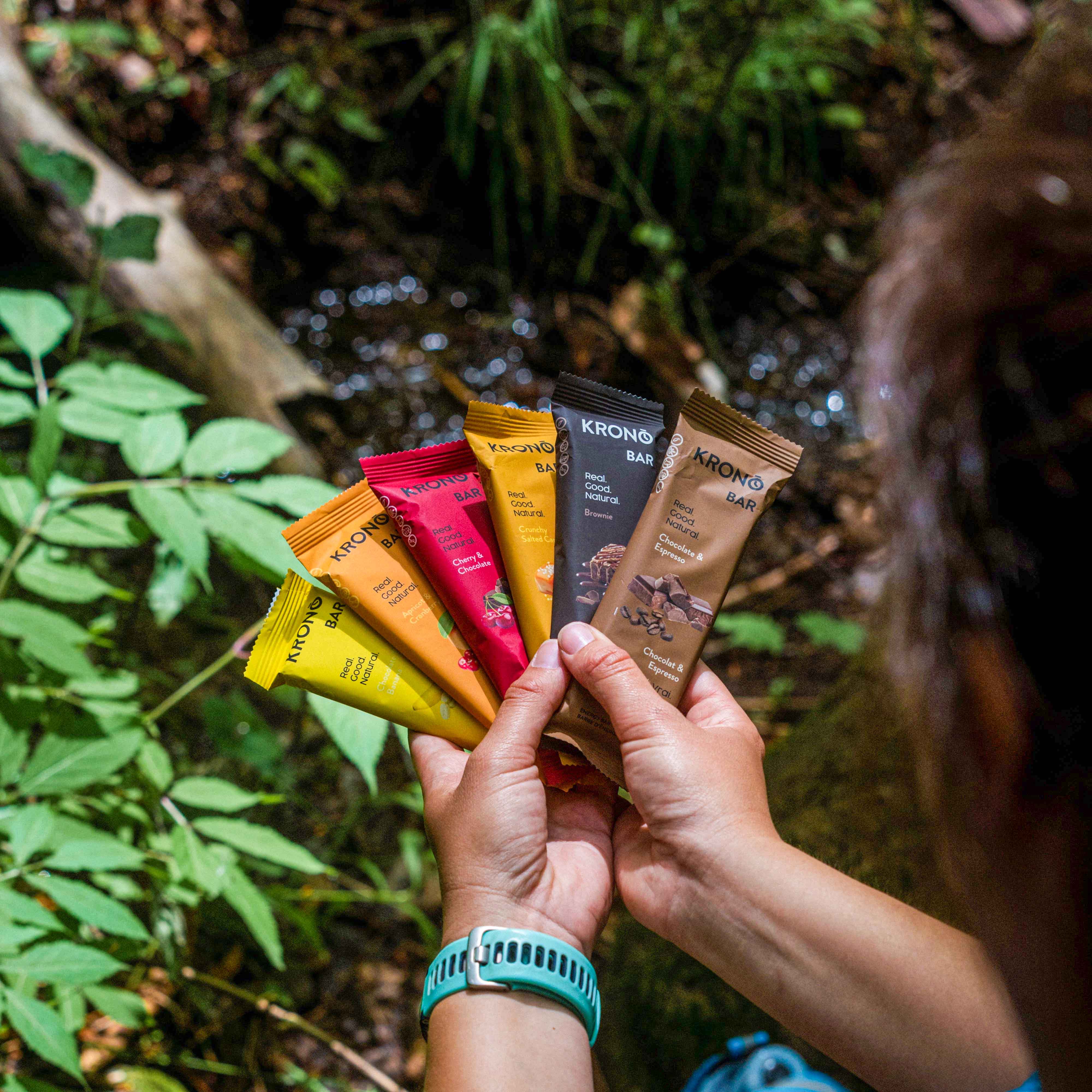 Hiker holding all five Krono Nutrition flavors of energy bars from the variety pack, on a trail