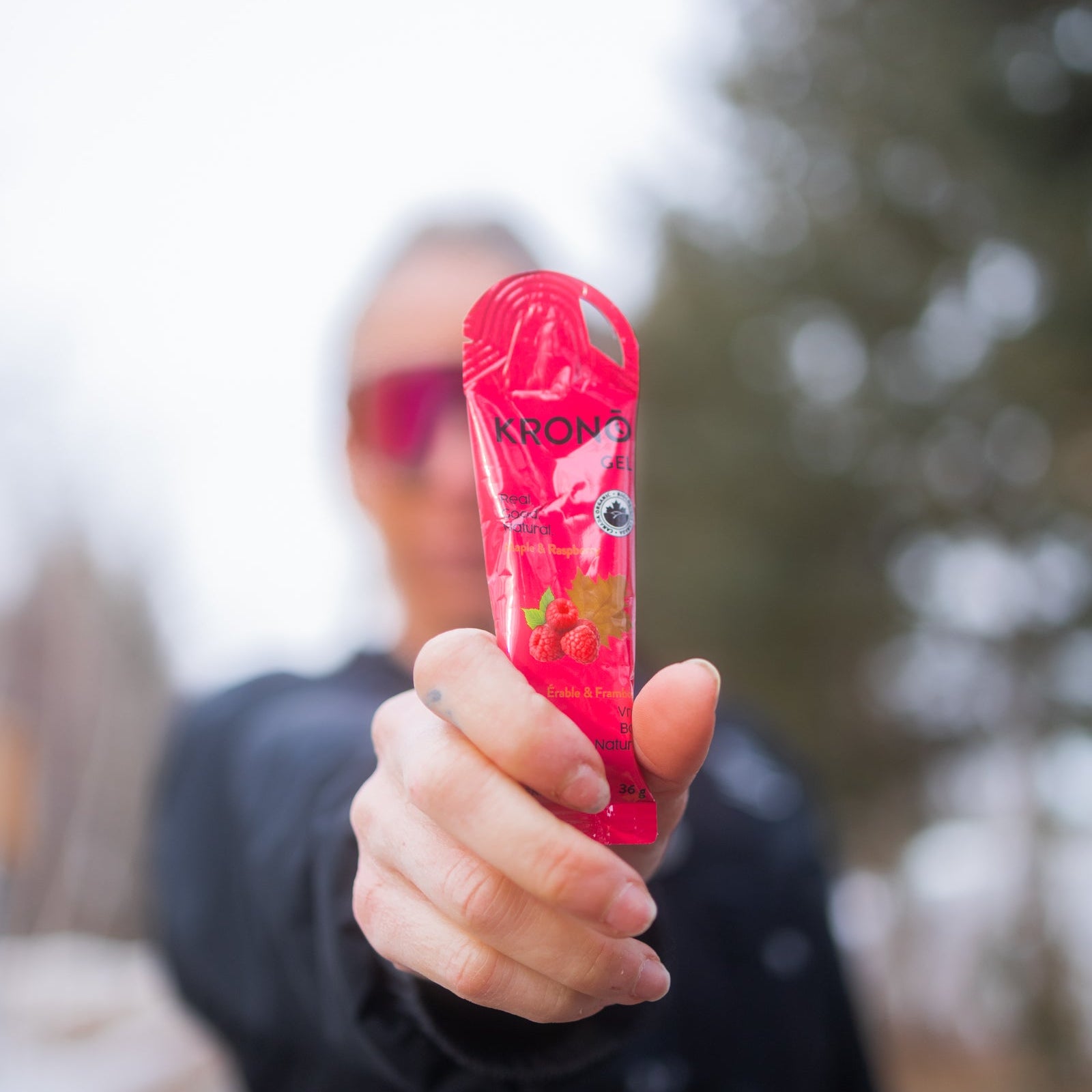 Close-up of a Krono Nutrition maple and raspberry energy gel being held by a person, with a blurry outdoors background