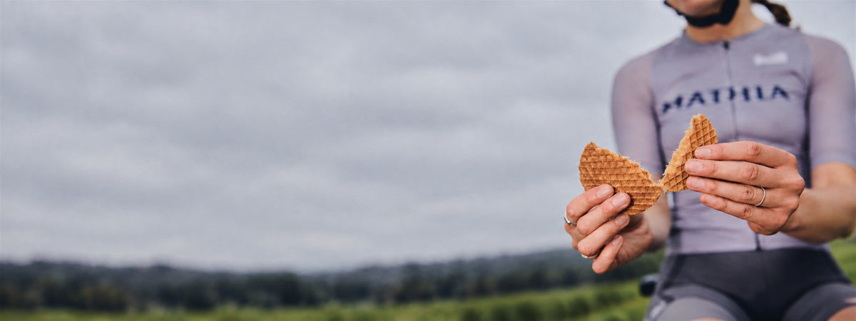 A cyclist in a gray jersey holds a waffle pieces outdoors against an overcast sky and green landscape.