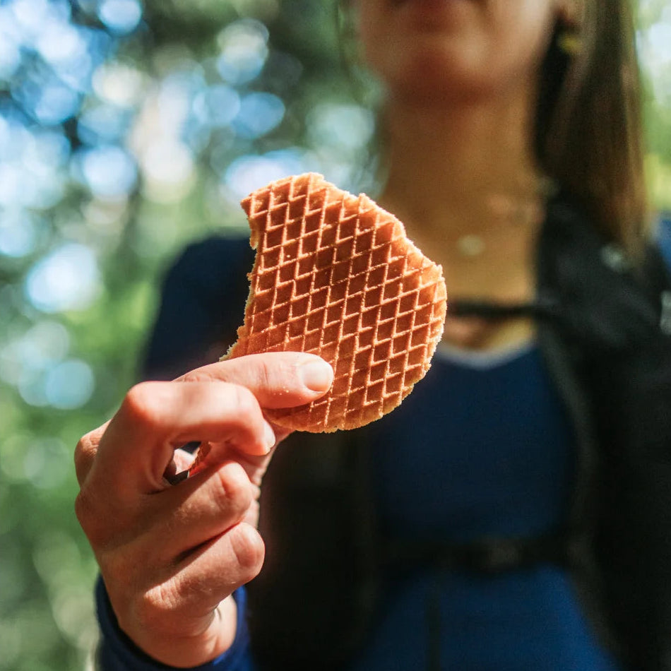 A person outdoors holds up a partially eaten waffle or waffle-like snack toward the camera, with a blurred green and blue natural background.