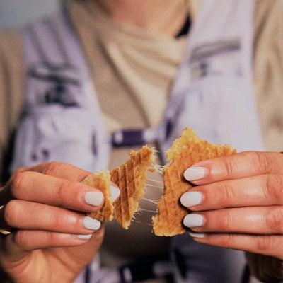 A close-up of a person wearing a light purple utility vest over a beige shirt, holding a waffle that is being pulled apart to show the stretchy fabric.