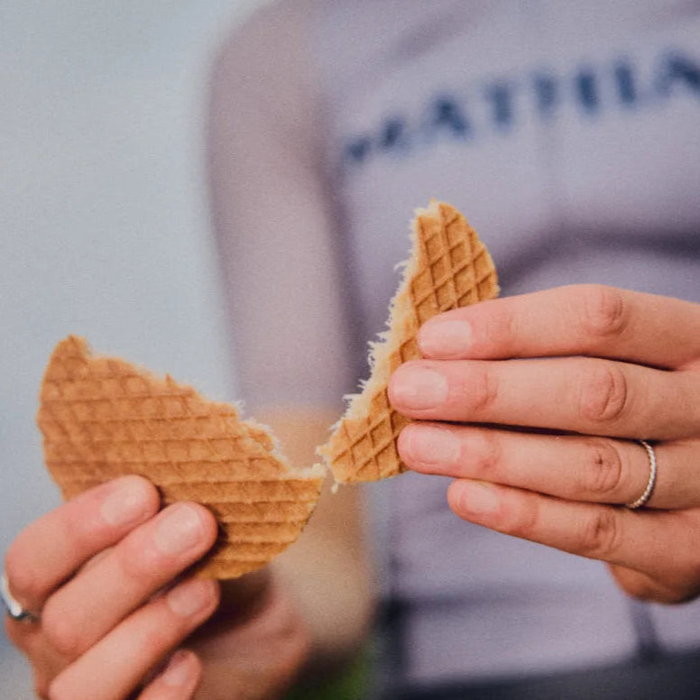 A close-up lifestyle shot of a person holding two halves of a broken waffle, with their hands in sharp focus and a blurred cyclist in a gray jersey in the background.