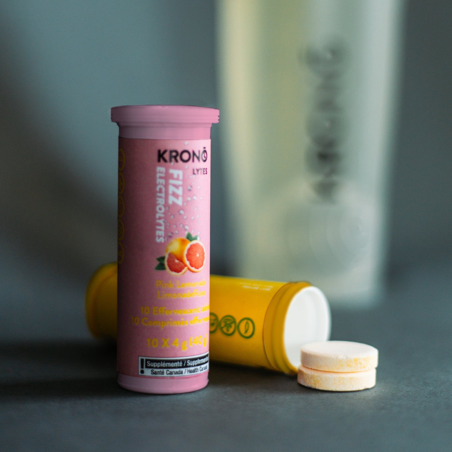 Standing Krono Nutrition pink lemonade effervescent tablets tube next to a laying lemon and lime tube and two tablets, blurry water bottle in the background