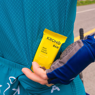 Cyclist taking out of his cycling jersey's back pocket a Krono Nutrition chocolate and banana protein bar with a road in the background