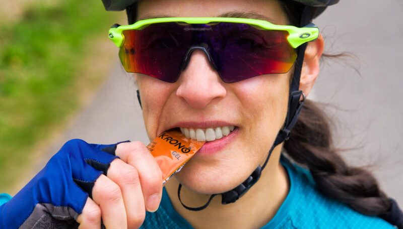 Jeune femme cycliste portant casque et lunettes qui ouvre avec ses dents un gel énergétique à l'érable Krono Nutrition