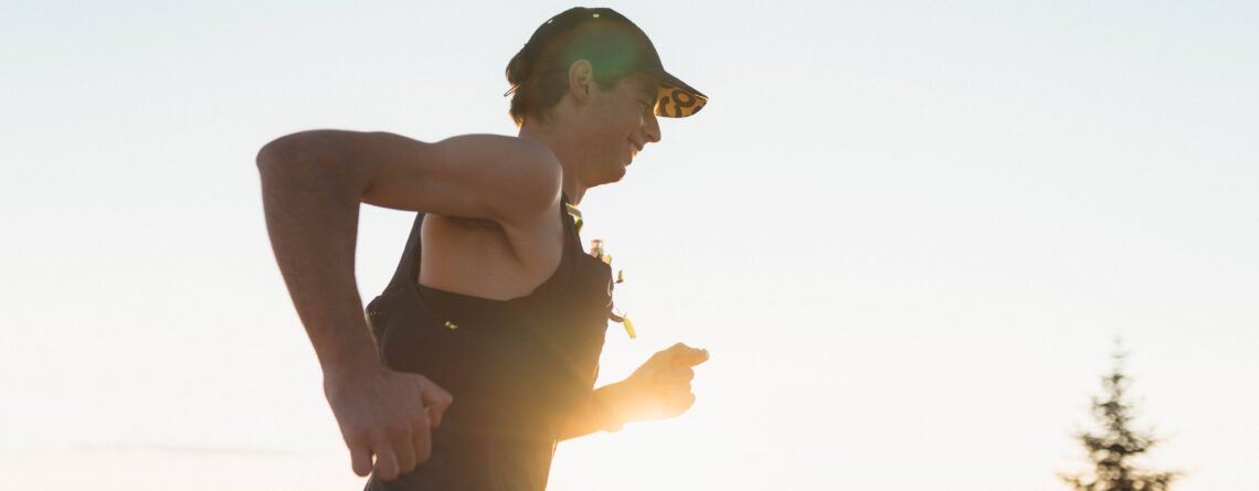 Coureur en action par une journée ensoleillée, portant une casquette et une veste d'hydration, dans un paysage montagneux